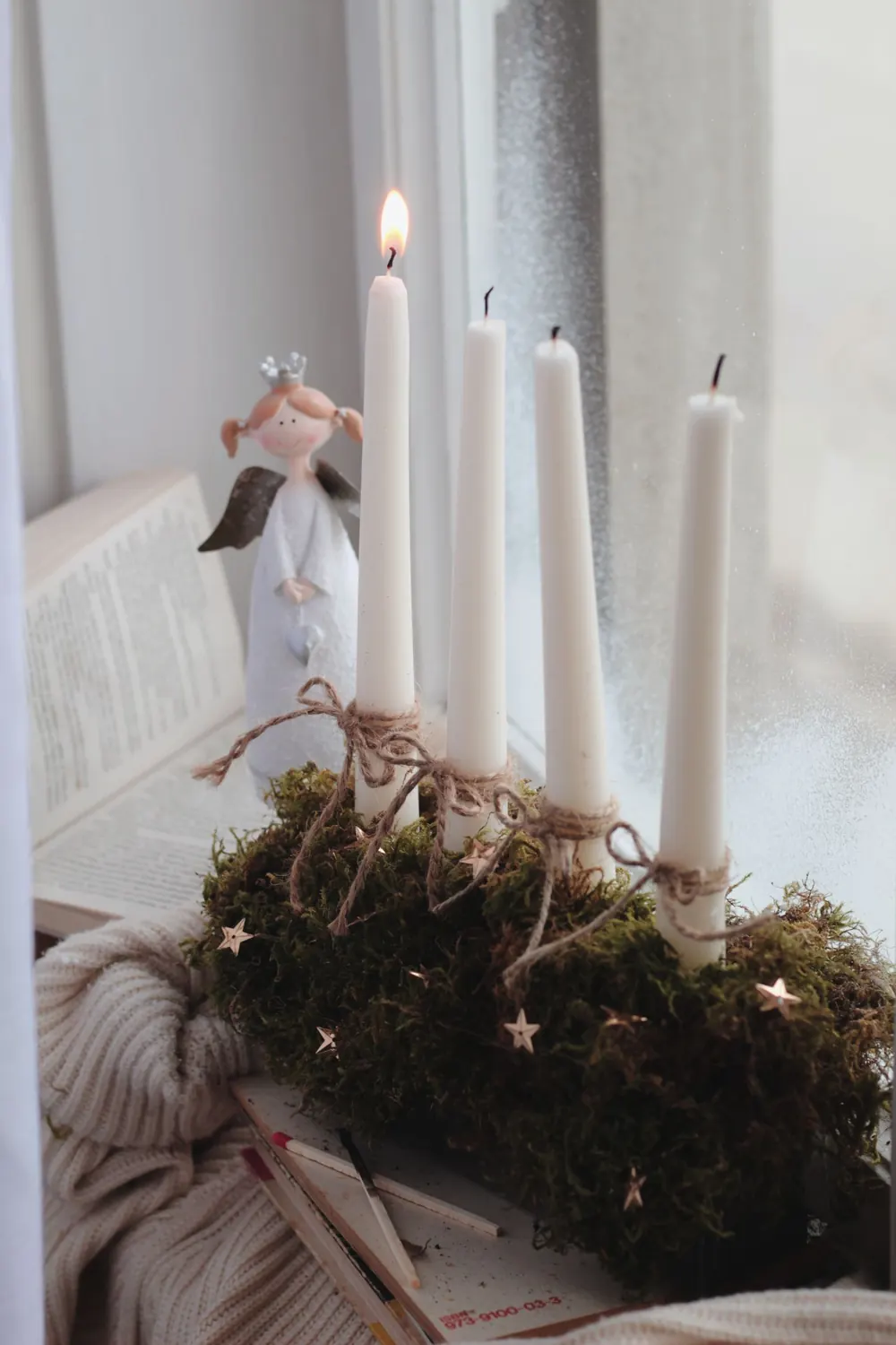 A serene still life featuring candles and an angel figurine by a window, evoking a cozy winter ambiance.